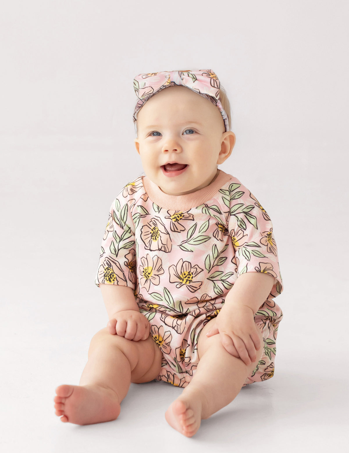 A smiling baby wears the Mabel and Honey Quinn Short Sleeve Romper, a soft, breathable pink floral outfit with a matching headband, sitting comfortably on a light surface against a plain background.