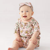 A smiling baby wears the Mabel and Honey Quinn Short Sleeve Romper, a soft, breathable pink floral outfit with a matching headband, sitting comfortably on a light surface against a plain background.