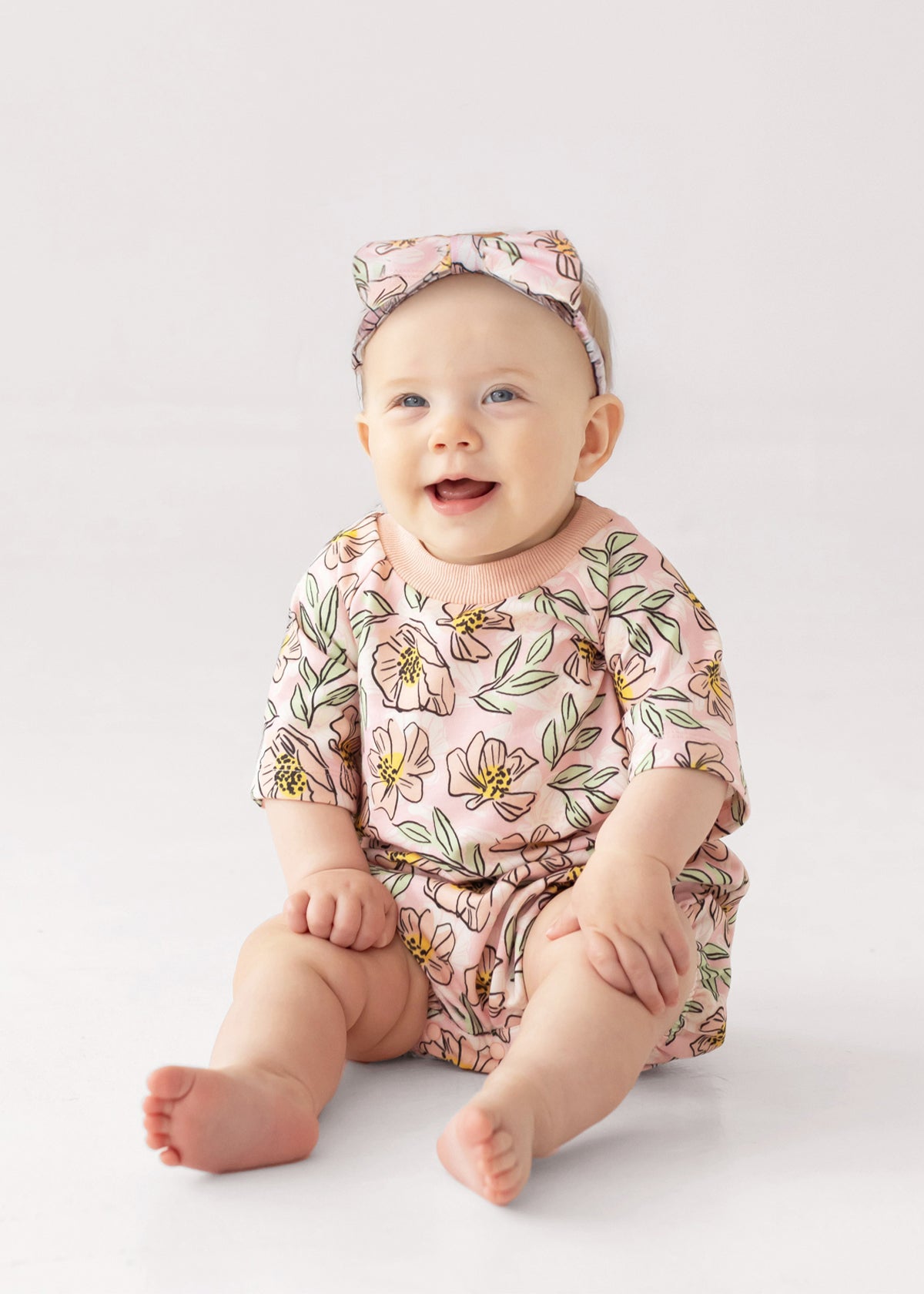 A smiling baby wears the Mabel and Honey Quinn Short Sleeve Romper, a soft, breathable pink floral outfit with a matching headband, sitting comfortably on a light surface against a plain background.
