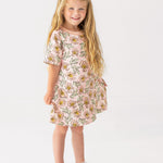 A young girl with long blonde hair smiles in the Mabel and Honey Quinn Short Sleeve Dress—a breathable, pink floral dress paired with white sandals—posing against a plain white background, perfect for everyday wear.