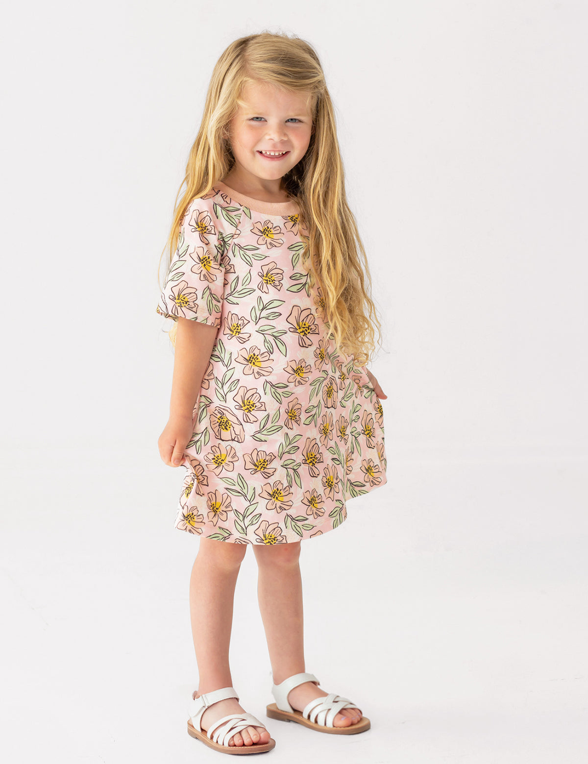A young girl with long blonde hair smiles in the Mabel and Honey Quinn Short Sleeve Dress—a breathable, pink floral dress paired with white sandals—posing against a plain white background, perfect for everyday wear.