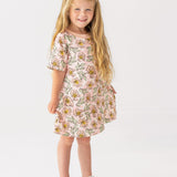 A young girl with long blonde hair smiles in the Mabel and Honey Quinn Short Sleeve Dress—a breathable, pink floral dress paired with white sandals—posing against a plain white background, perfect for everyday wear.