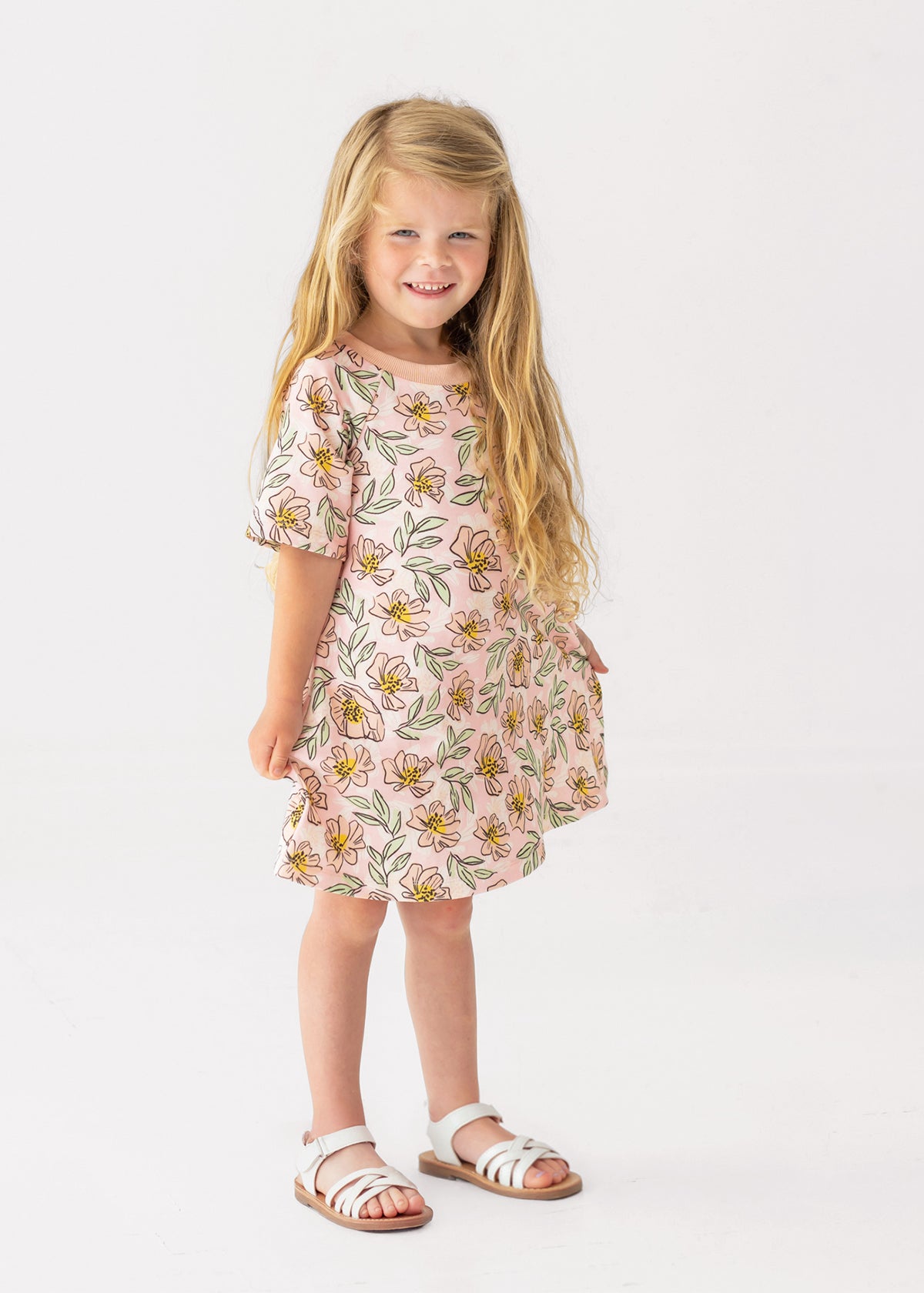A young girl with long blonde hair smiles in the Mabel and Honey Quinn Short Sleeve Dress—a breathable, pink floral dress paired with white sandals—posing against a plain white background, perfect for everyday wear.