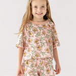 A young girl with long, light brown hair smiles at the camera wearing the Mabel and Honey Quinn Short Sleeve Two Piece Set—breathable shorts and a top adorned with pink flowers and green leaves—against a plain white background.