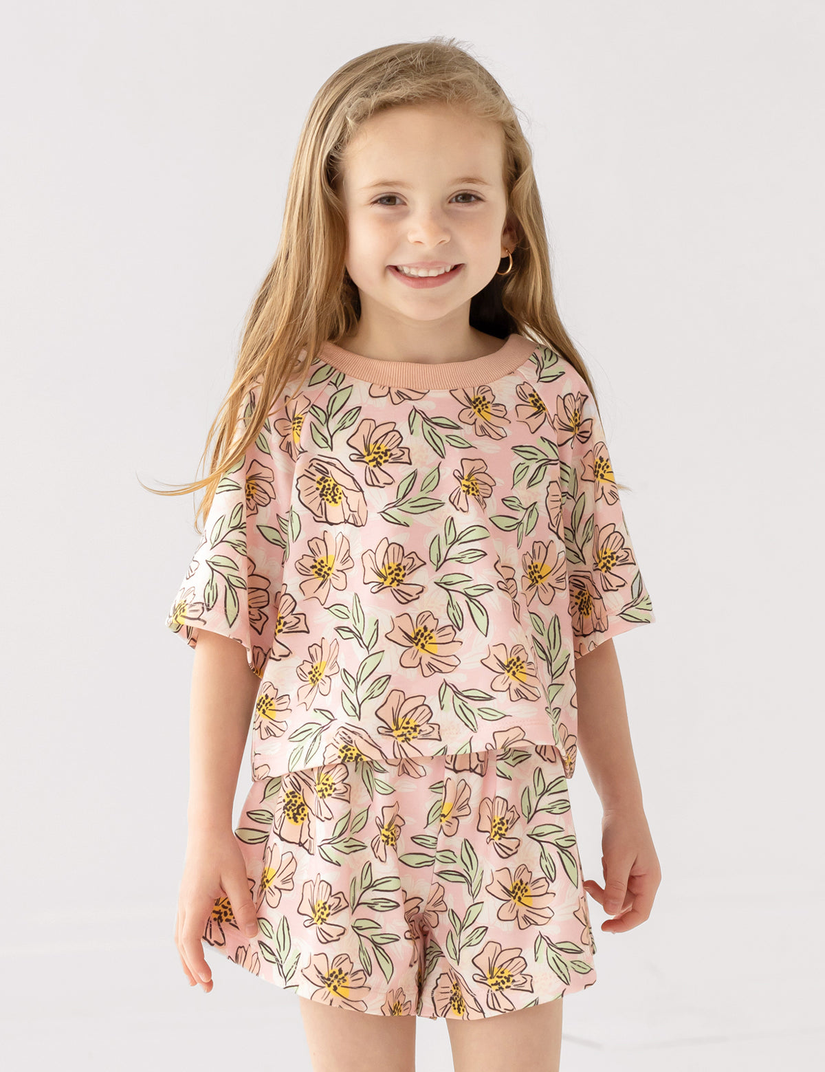 A young girl with long, light brown hair smiles at the camera wearing the Mabel and Honey Quinn Short Sleeve Two Piece Set—breathable shorts and a top adorned with pink flowers and green leaves—against a plain white background.