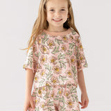 A young girl with long, light brown hair smiles at the camera wearing the Mabel and Honey Quinn Short Sleeve Two Piece Set—breathable shorts and a top adorned with pink flowers and green leaves—against a plain white background.