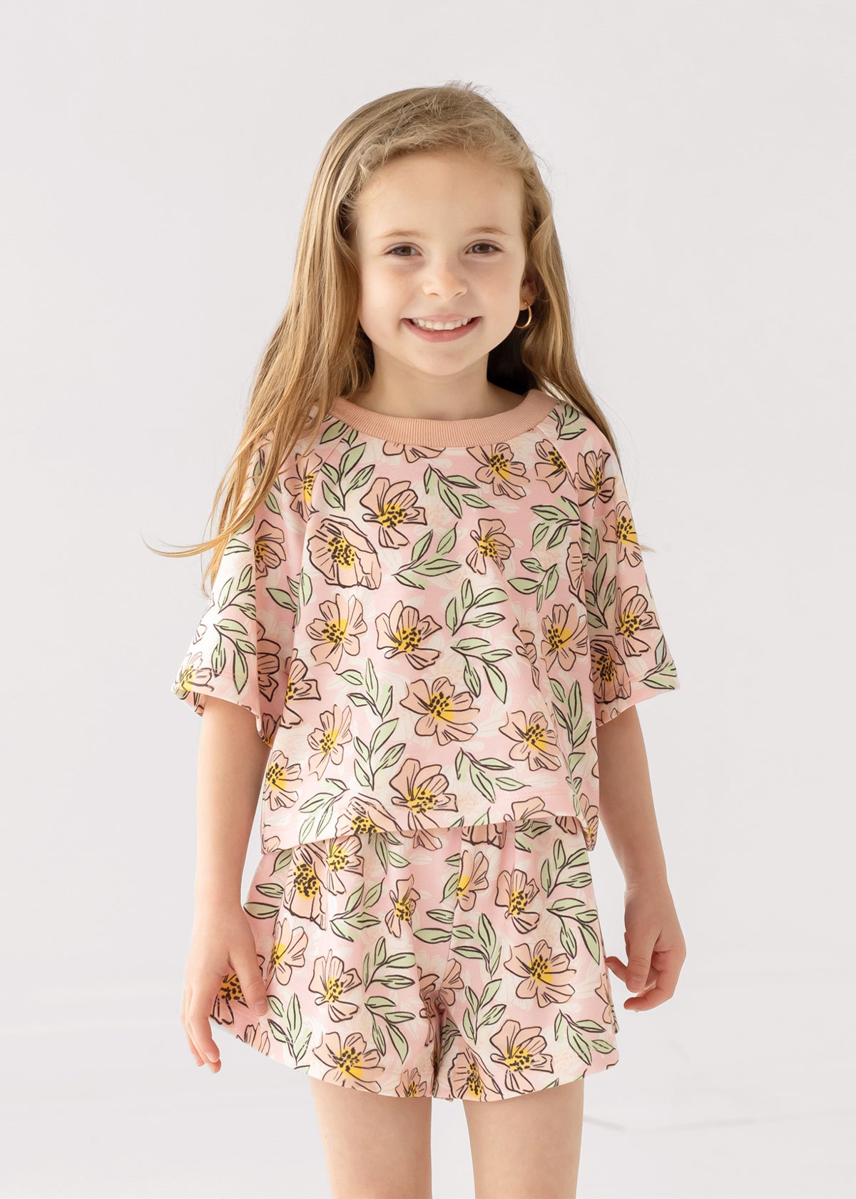 A young girl with long, light brown hair smiles at the camera wearing the Mabel and Honey Quinn Short Sleeve Two Piece Set—breathable shorts and a top adorned with pink flowers and green leaves—against a plain white background.