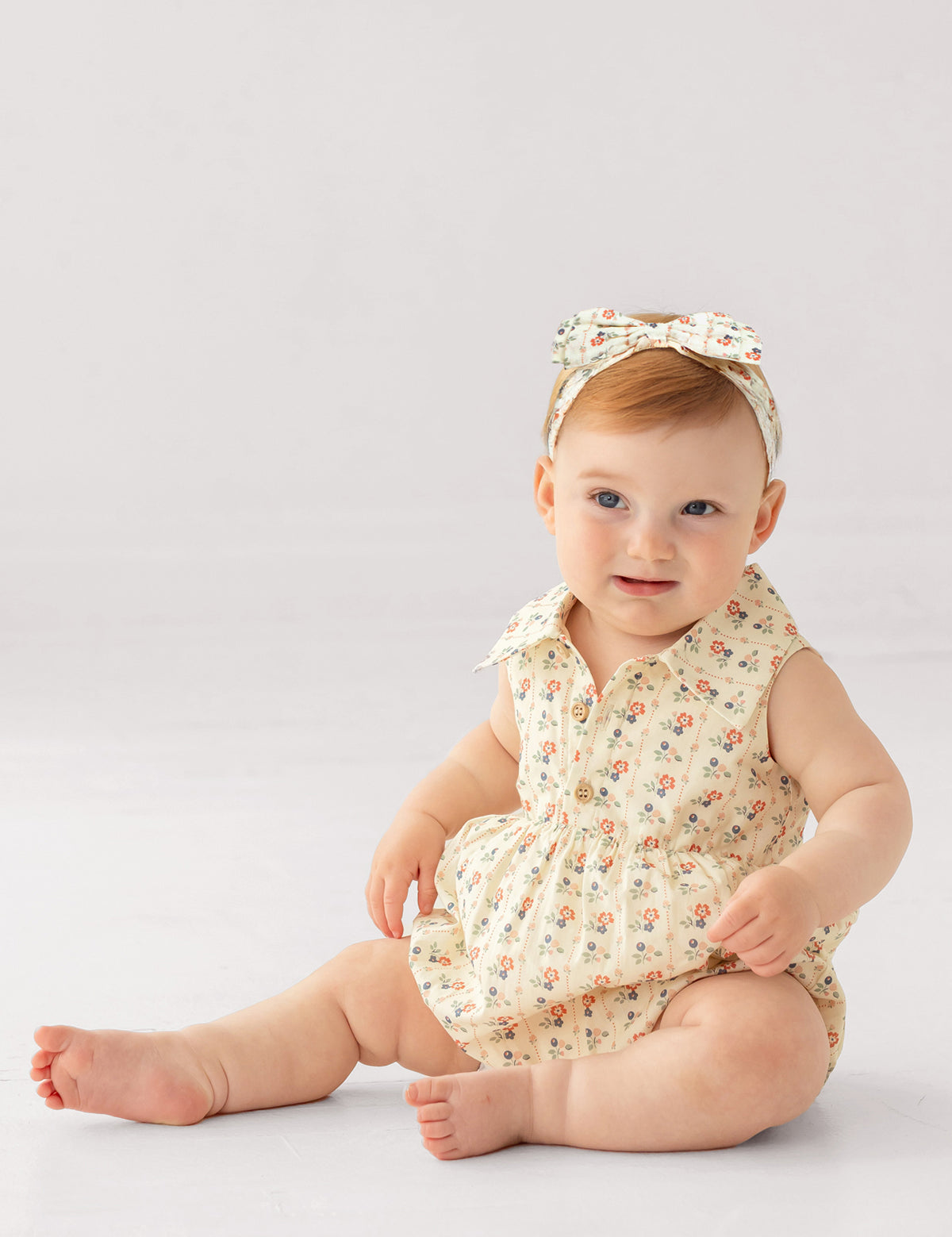 A light-skinned baby with short light brown hair sits on the floor wearing the Adelaide Sleeveless Romper by Mabel and Honey, featuring a vintage floral print, wooden buttons, and a matching bow headband, against a plain white background.
