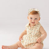 A light-skinned baby with short light brown hair sits on the floor wearing the Adelaide Sleeveless Romper by Mabel and Honey, featuring a vintage floral print, wooden buttons, and a matching bow headband, against a plain white background.