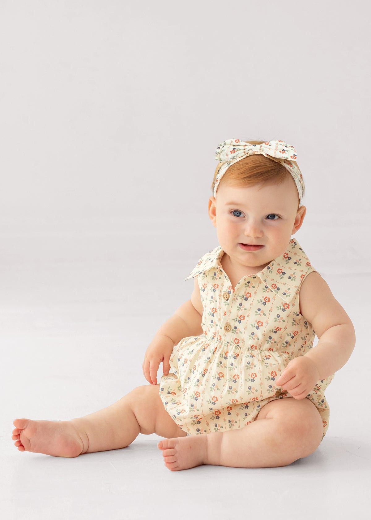 A light-skinned baby with short light brown hair sits on the floor wearing the Adelaide Sleeveless Romper by Mabel and Honey, featuring a vintage floral print, wooden buttons, and a matching bow headband, against a plain white background.