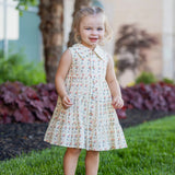 A young girl with light hair in pigtails stands on green grass, wearing the Mabel and Honey Adelaide Sleeveless Dress. She smiles softly, with trees, bushes, and a building blurred in the background.