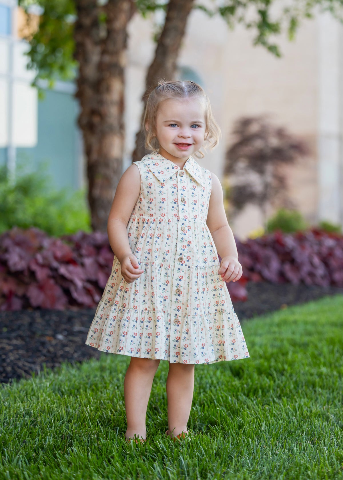 A young girl with light hair in pigtails stands on green grass, wearing the Mabel and Honey Adelaide Sleeveless Dress. She smiles softly, with trees, bushes, and a building blurred in the background.