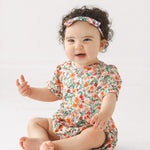 A smiling baby with curly hair sits barefoot on the floor, wearing the Mabel and Honey Florentine Short Gathered Sleeve Romper with a floral print and matching headband, set against a plain white background.