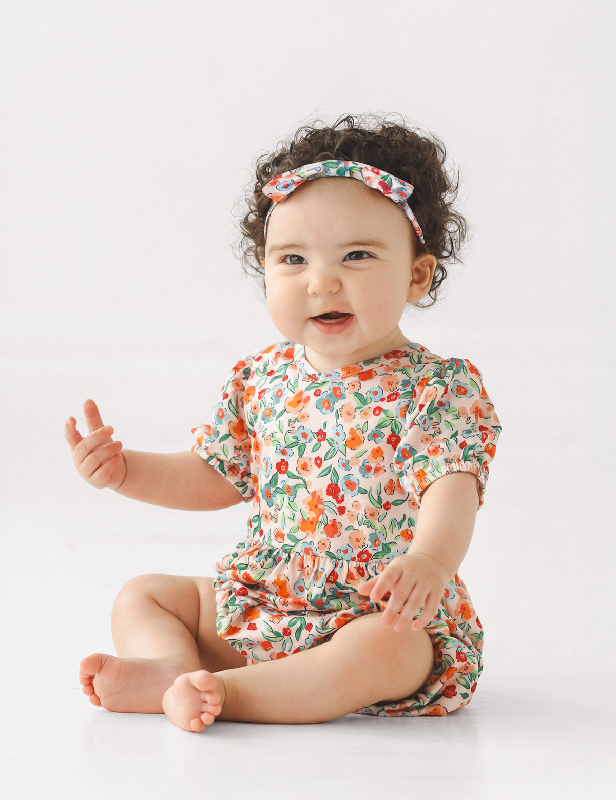 A smiling baby with curly hair sits barefoot on the floor, wearing the Mabel and Honey Florentine Short Gathered Sleeve Romper with a floral print and matching headband, set against a plain white background.