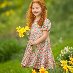 A young girl with curly red hair stands outdoors on green grass, smiling and holding yellow daffodils while wearing the Mabel and Honey Florentine Short Gathered Sleeve Dress amid blooming flowers.