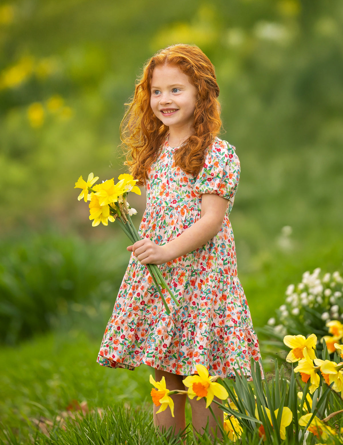 A young girl with curly red hair stands outdoors on green grass, smiling and holding yellow daffodils while wearing the Mabel and Honey Florentine Short Gathered Sleeve Dress amid blooming flowers.