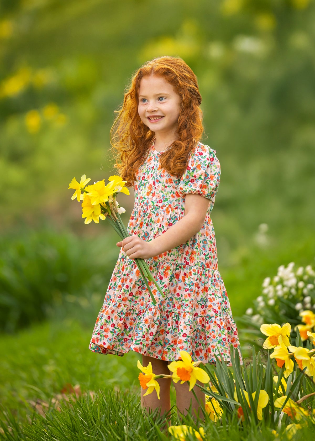 A young girl with curly red hair stands outdoors on green grass, smiling and holding yellow daffodils while wearing the Mabel and Honey Florentine Short Gathered Sleeve Dress amid blooming flowers.