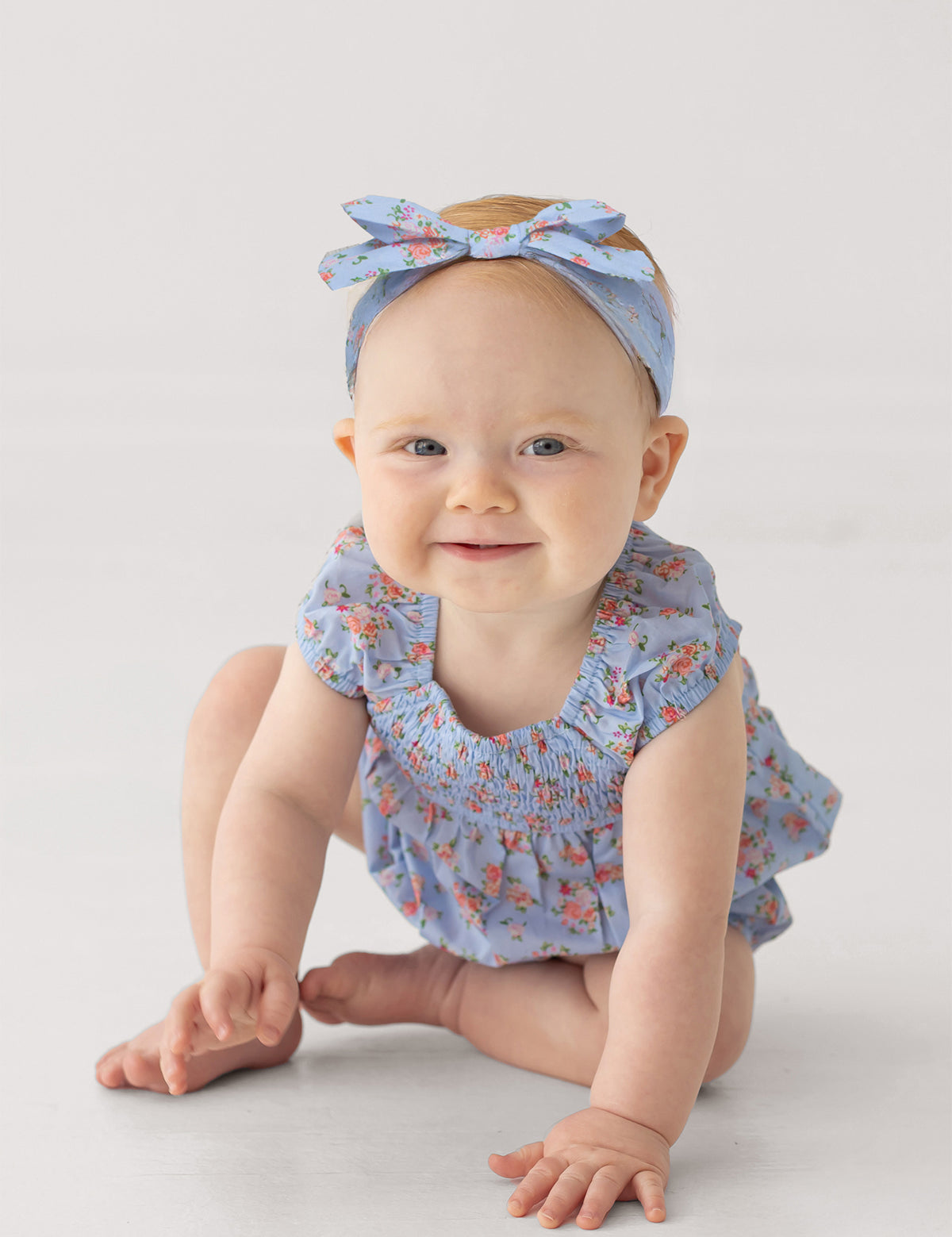 A baby sits on the floor, smiling softly against a light background, wearing Mabel and Honey’s Delia Sleeveless Romper in light blue with ruffled cap sleeves and a matching headband.
