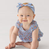 A baby sits on the floor, smiling softly against a light background, wearing Mabel and Honey’s Delia Sleeveless Romper in light blue with ruffled cap sleeves and a matching headband.