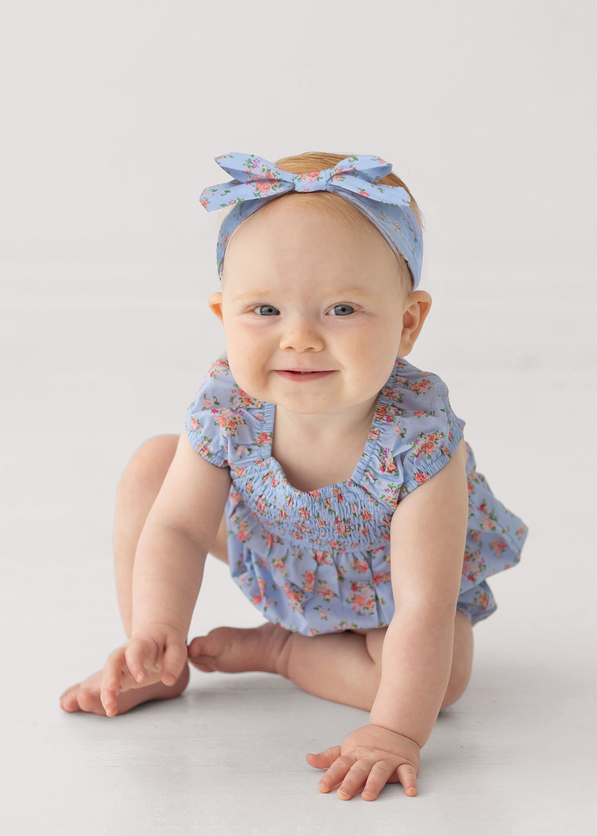 A baby sits on the floor, smiling softly against a light background, wearing Mabel and Honey’s Delia Sleeveless Romper in light blue with ruffled cap sleeves and a matching headband.