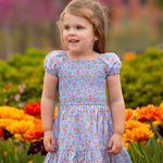 A young girl with light brown hair smiles while wearing the Mabel and Honey Delia Cap Sleeve Dress. She stands among orange, yellow, and pink flowers, with green bushes and blurred trees in the background.