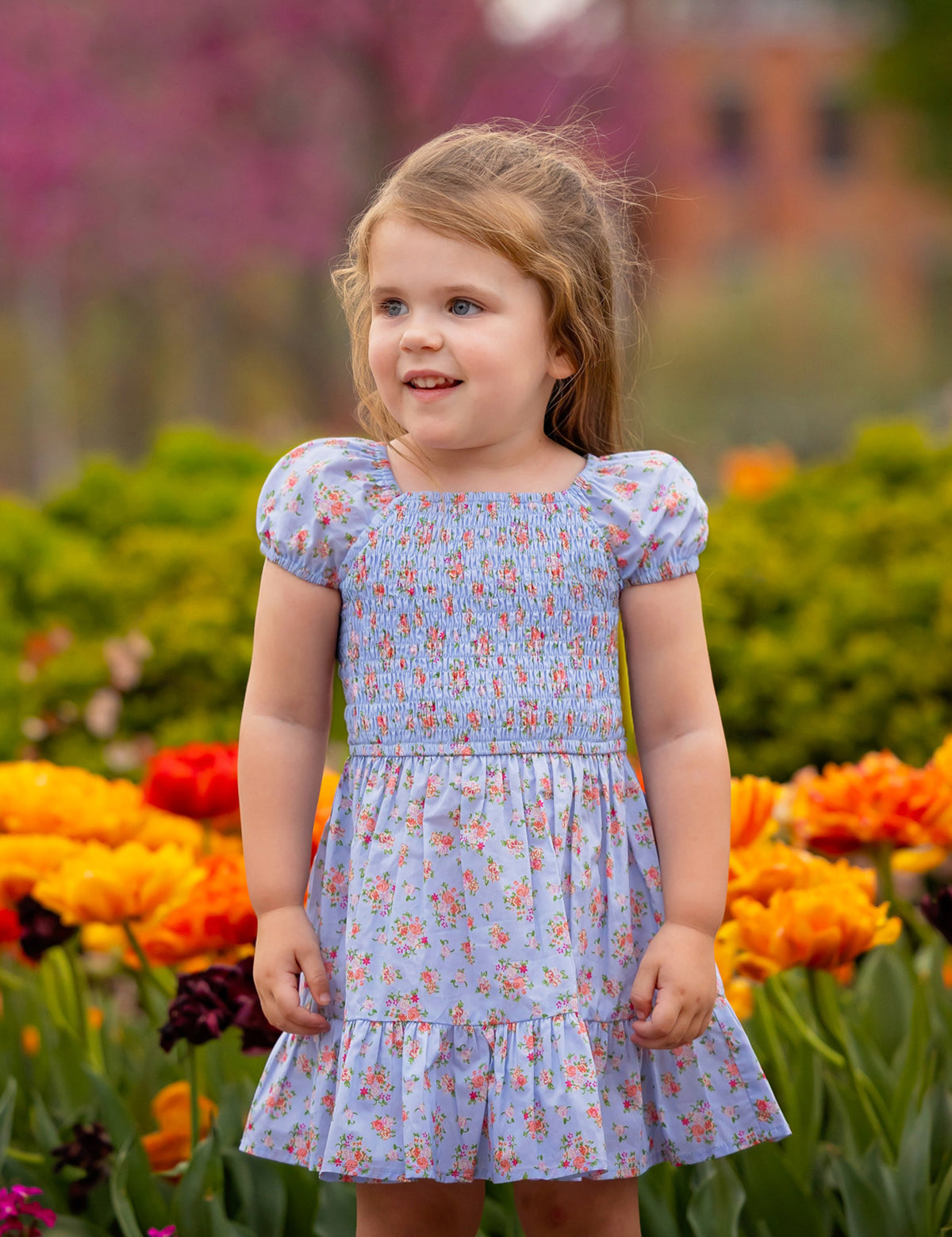 A young girl with light brown hair smiles while wearing the Mabel and Honey Delia Cap Sleeve Dress. She stands among orange, yellow, and pink flowers, with green bushes and blurred trees in the background.