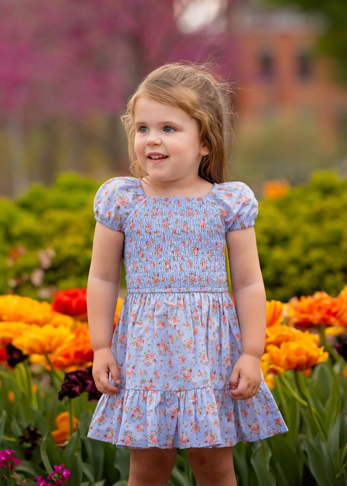 A young girl with light brown hair smiles while wearing the Mabel and Honey Delia Cap Sleeve Dress. She stands among orange, yellow, and pink flowers, with green bushes and blurred trees in the background.