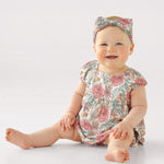 A smiling baby sits on the floor wearing the Mabel and Honey April Cap Sleeve Romper with a square neckline and matching floral headband, set against a plain white background.