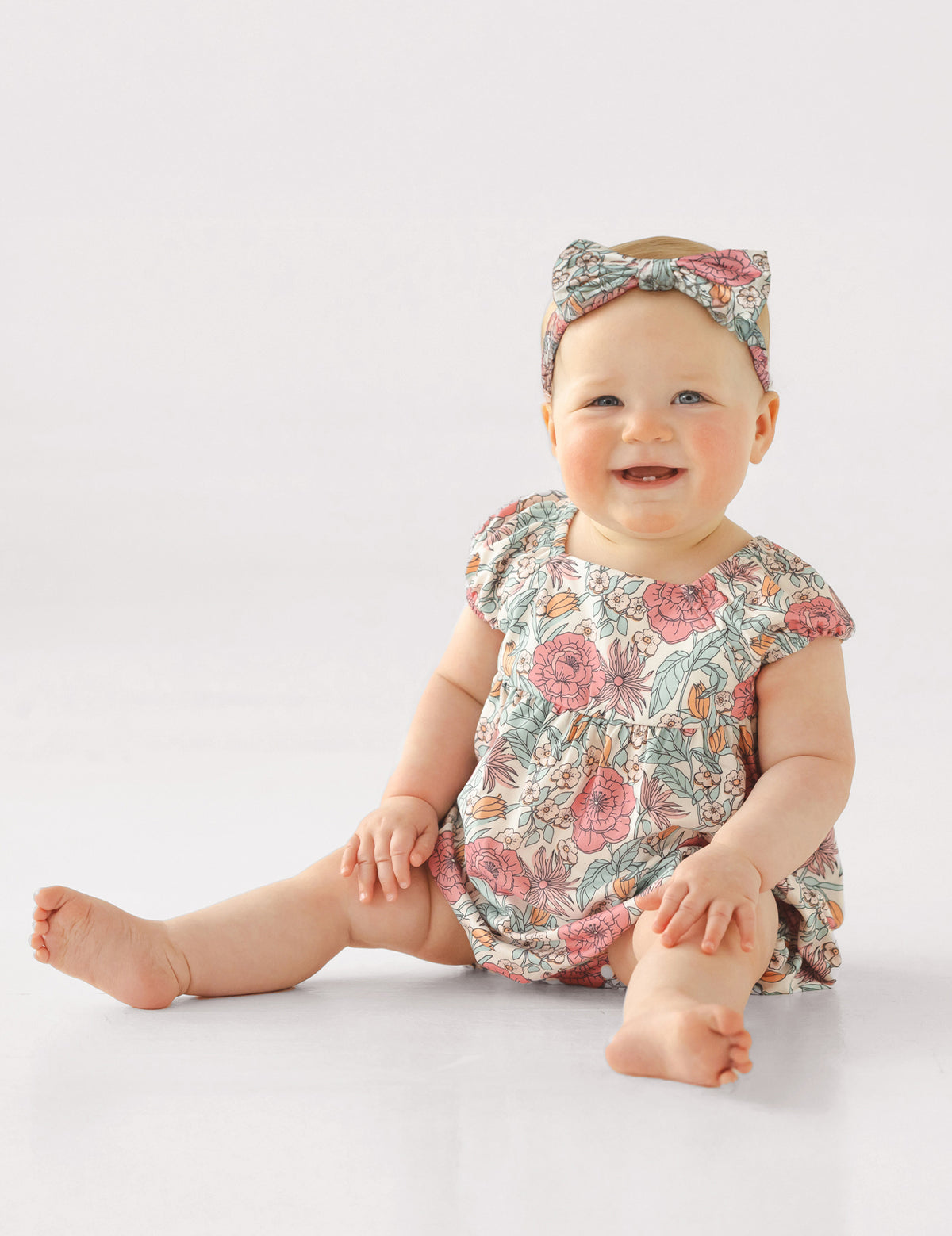 A smiling baby sits on the floor wearing the Mabel and Honey April Cap Sleeve Romper with a square neckline and matching floral headband, set against a plain white background.