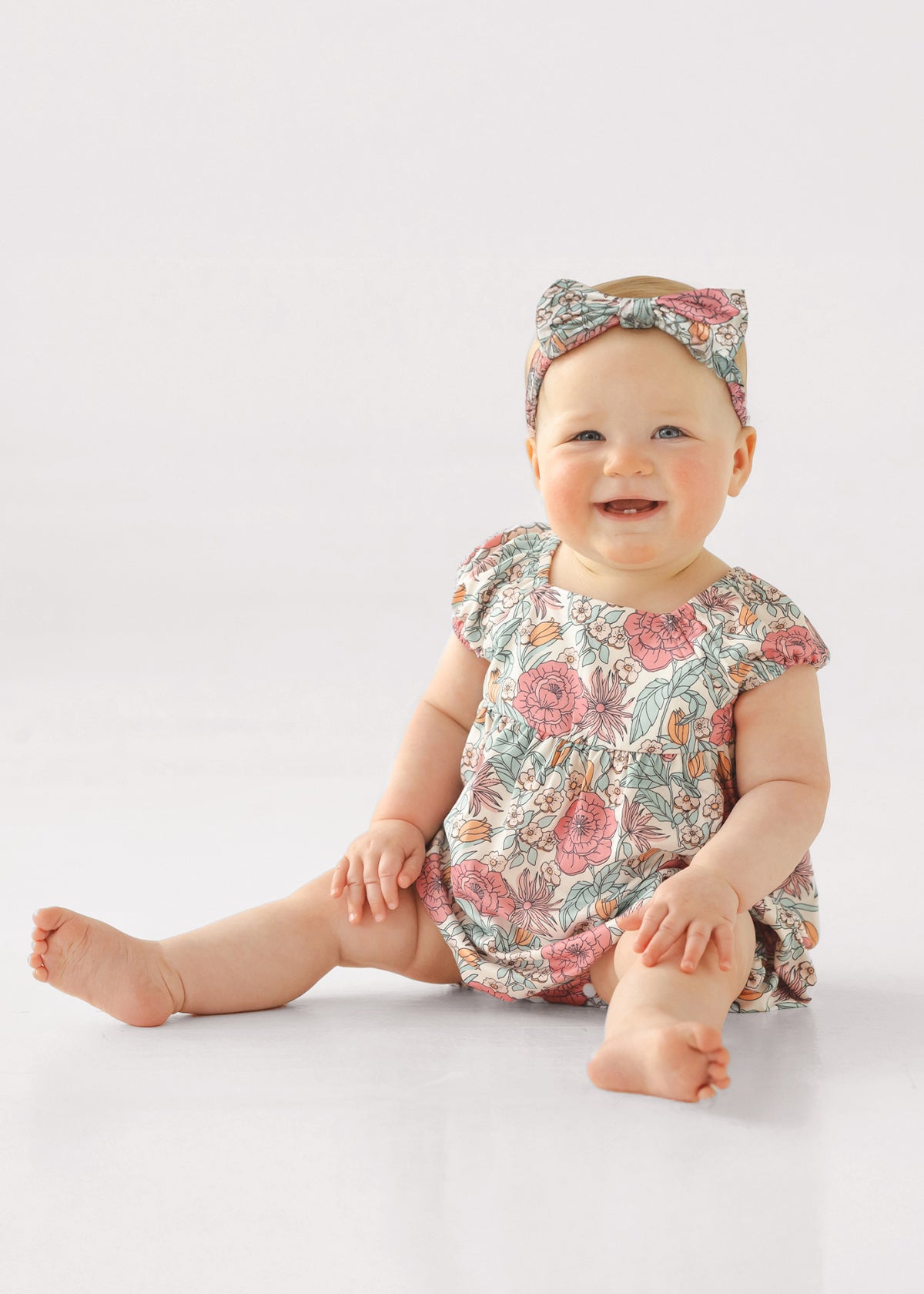A smiling baby sits on the floor wearing the Mabel and Honey April Cap Sleeve Romper with a square neckline and matching floral headband, set against a plain white background.