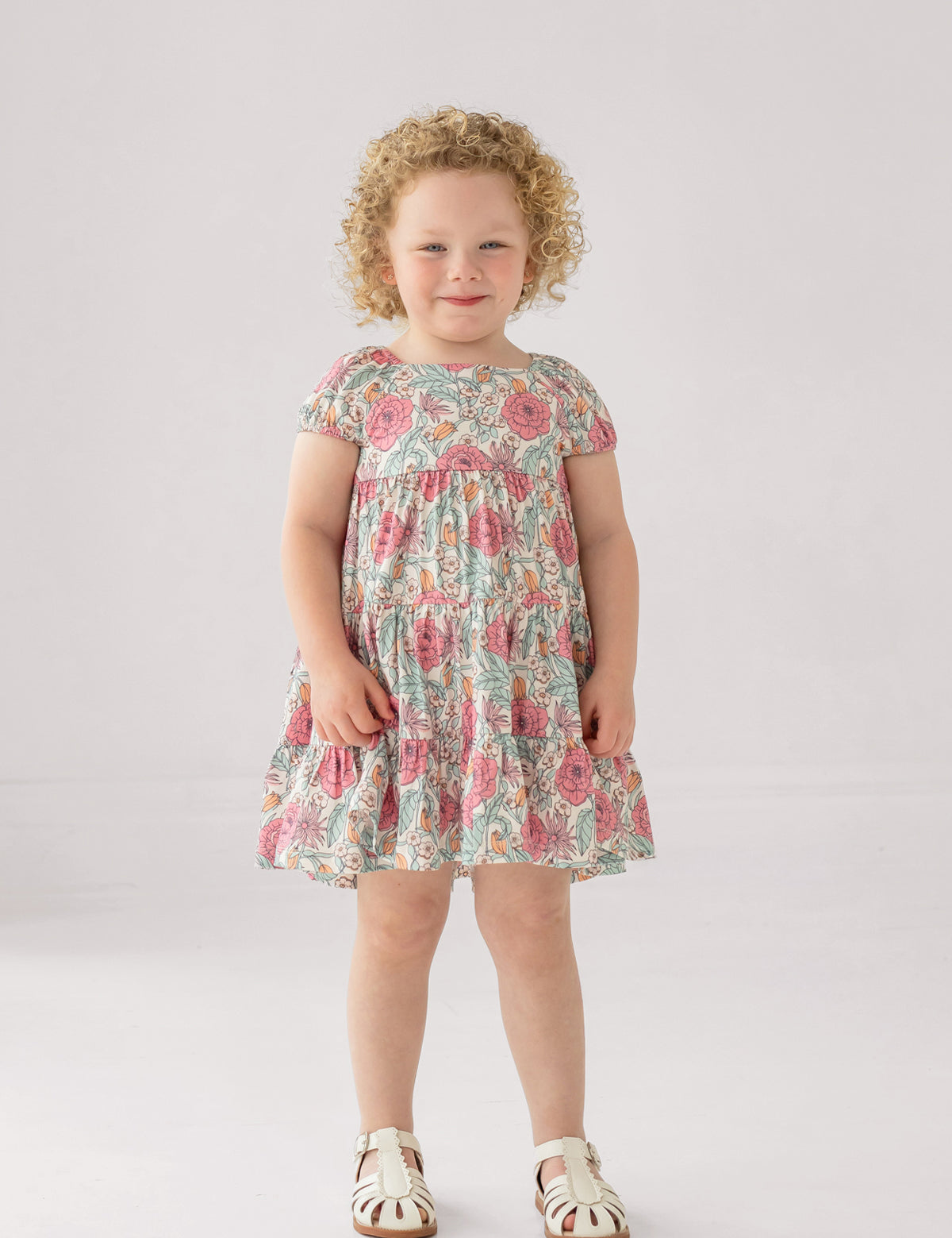 A girl wears the Mabel and Honey April Cap Sleeve Dress, featuring a floral print and a tiered skirt.