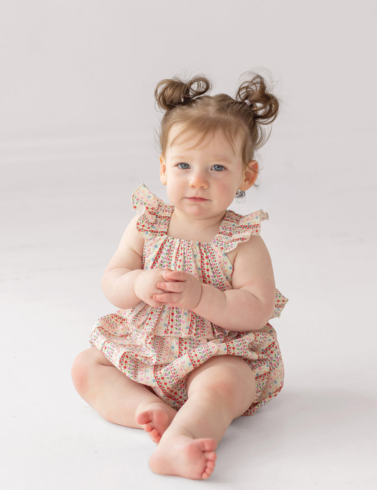 A light-skinned baby with brown hair in pigtails sits barefoot on a white surface, wearing the Bella Reina Sleeveless Romper by Mabel and Honey, and gazes calmly past the camera.