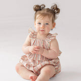 A light-skinned baby with brown hair in pigtails sits barefoot on a white surface, wearing the Bella Reina Sleeveless Romper by Mabel and Honey, and gazes calmly past the camera.