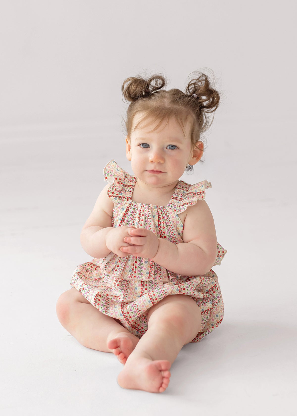 A light-skinned baby with brown hair in pigtails sits barefoot on a white surface, wearing the Bella Reina Sleeveless Romper by Mabel and Honey, and gazes calmly past the camera.