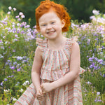A young child with curly red hair smiles in a field of purple and white wildflowers, wearing the Bella Reina Sleeveless Dress by Mabel and Honey—a patterned dress ideal for special occasions against a lush green backdrop.