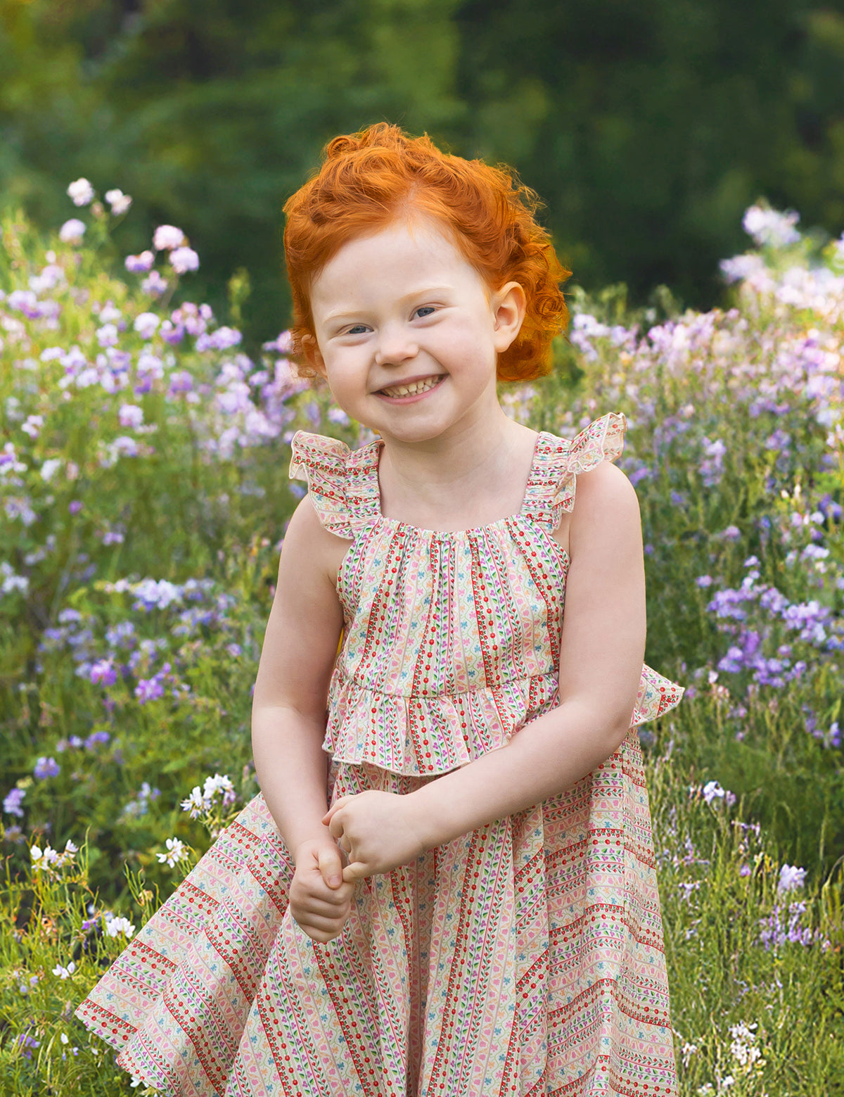 A young child with curly red hair smiles in a field of purple and white wildflowers, wearing the Bella Reina Sleeveless Dress by Mabel and Honey—a patterned dress ideal for special occasions against a lush green backdrop.