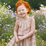 A young child with curly red hair smiles in a field of purple and white wildflowers, wearing the Bella Reina Sleeveless Dress by Mabel and Honey—a patterned dress ideal for special occasions against a lush green backdrop.