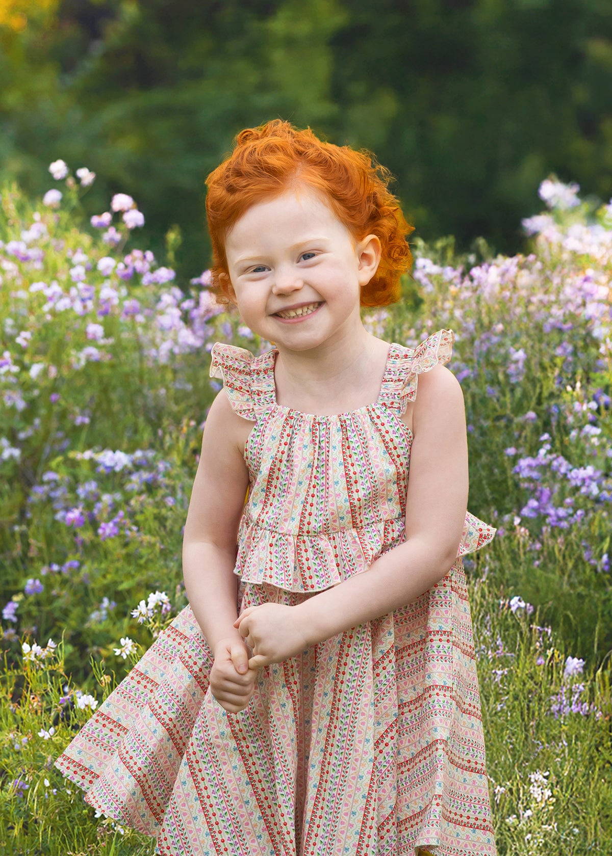 A young child with curly red hair smiles in a field of purple and white wildflowers, wearing the Bella Reina Sleeveless Dress by Mabel and Honey—a patterned dress ideal for special occasions against a lush green backdrop.