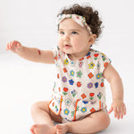 A baby with curly hair wears the Mabel and Honey Little Garden Parade Cap Sleeve Romper and matching headband, sitting on the floor with one arm raised against a plain white background.