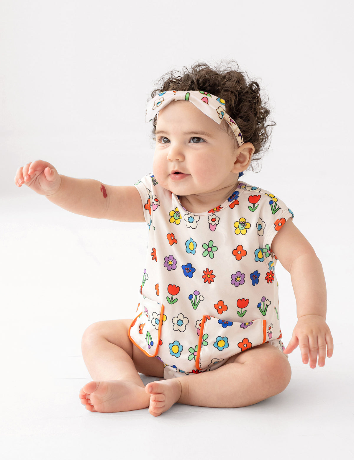 A baby with curly hair wears the Mabel and Honey Little Garden Parade Cap Sleeve Romper and matching headband, sitting on the floor with one arm raised against a plain white background.