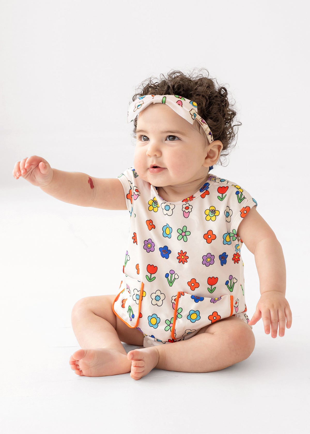 A baby with curly hair wears the Mabel and Honey Little Garden Parade Cap Sleeve Romper and matching headband, sitting on the floor with one arm raised against a plain white background.