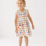 A young girl with blond hair in a top bun smiles while wearing the Mabel and Honey Little Garden Parade Cap Sleeve Dress, a white floral dress with colorful patterns and white sandals, standing against a plain white background.
