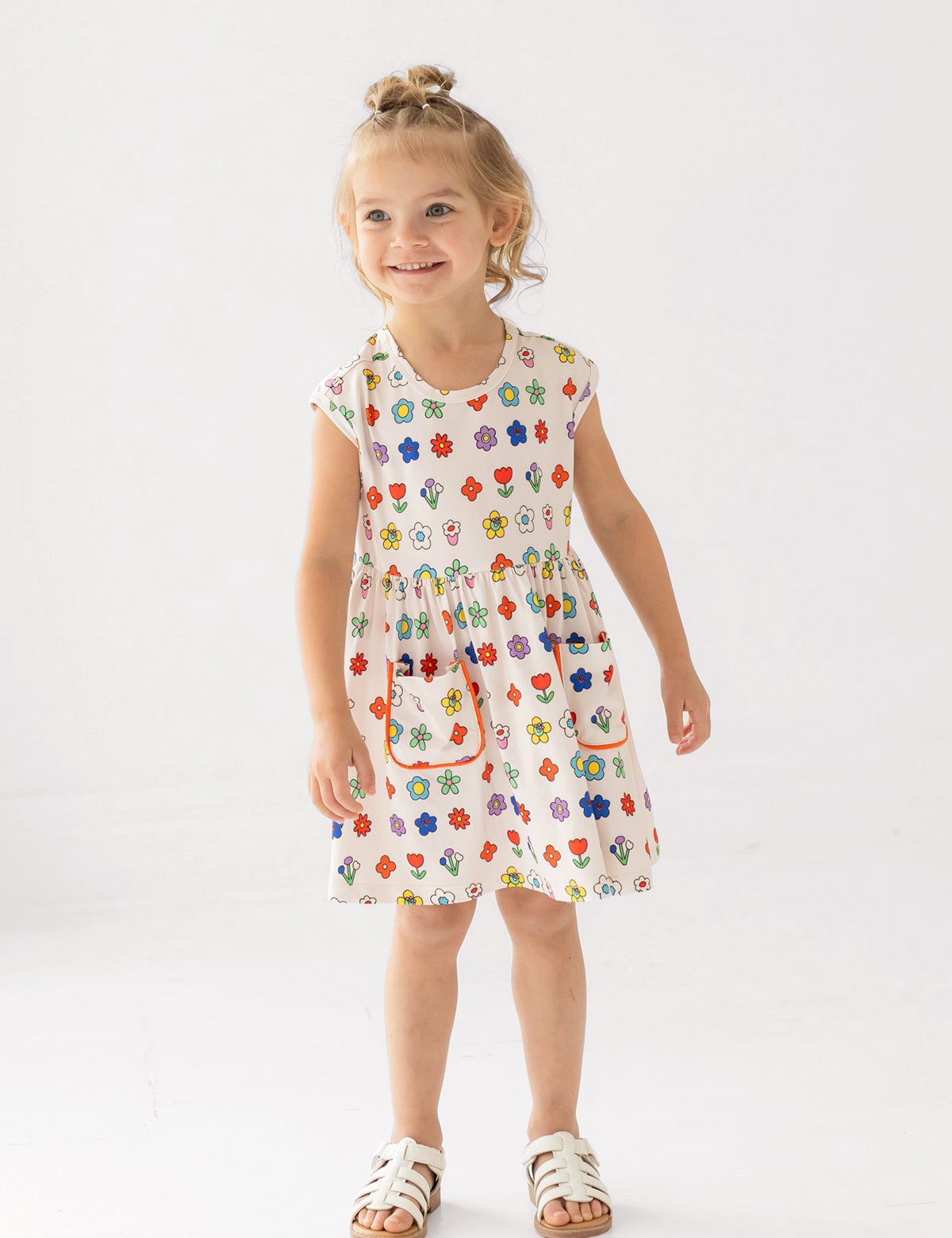 A young girl with blond hair in a top bun smiles while wearing the Mabel and Honey Little Garden Parade Cap Sleeve Dress, a white floral dress with colorful patterns and white sandals, standing against a plain white background.