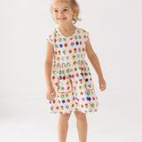A young girl with blond hair in a top bun smiles while wearing the Mabel and Honey Little Garden Parade Cap Sleeve Dress, a white floral dress with colorful patterns and white sandals, standing against a plain white background.