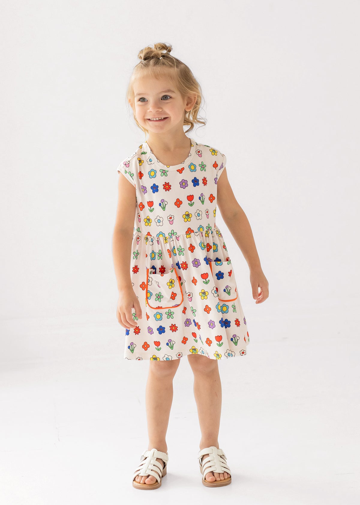 A young girl with blond hair in a top bun smiles while wearing the Mabel and Honey Little Garden Parade Cap Sleeve Dress, a white floral dress with colorful patterns and white sandals, standing against a plain white background.