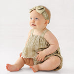 A light-skinned, blue-eyed baby wears the Mabel and Honey Espresso Sleeveless Romper with a matching headband and bow, sitting on the floor against a plain background and looking slightly upward.