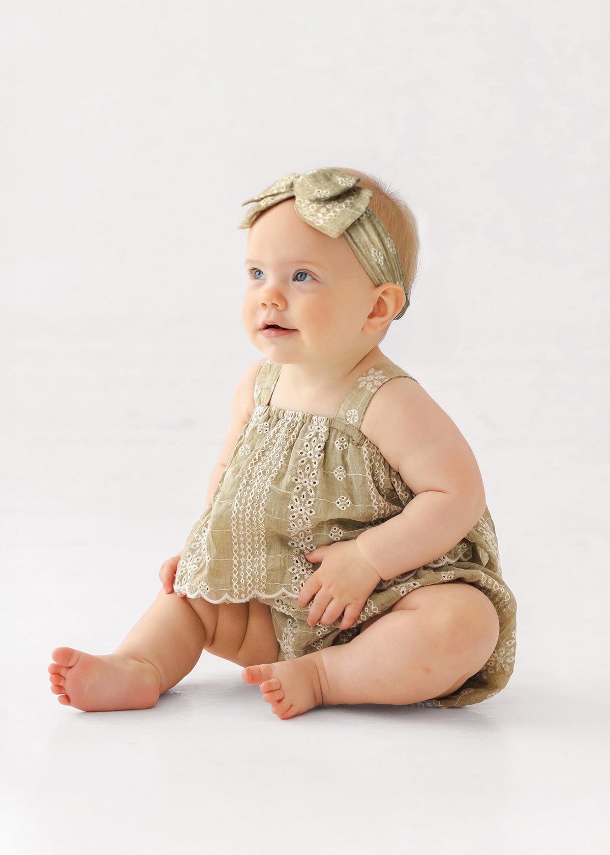 A light-skinned, blue-eyed baby wears the Mabel and Honey Espresso Sleeveless Romper with a matching headband and bow, sitting on the floor against a plain background and looking slightly upward.