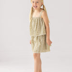 A young girl with blonde pigtails smiles in a Mabel and Honey Espresso Sleeveless Dress with embroidery and a scalloped hem, paired with white sandals. The background is plain and light-colored.