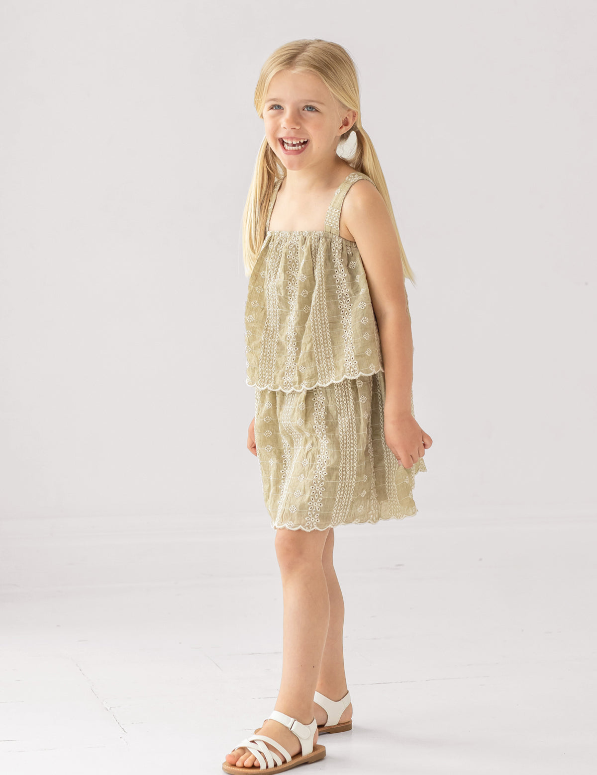 A young girl with blonde pigtails smiles in a Mabel and Honey Espresso Sleeveless Dress with embroidery and a scalloped hem, paired with white sandals. The background is plain and light-colored.