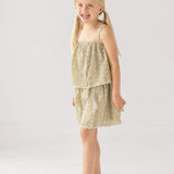 A young girl with blonde pigtails smiles in a Mabel and Honey Espresso Sleeveless Dress with embroidery and a scalloped hem, paired with white sandals. The background is plain and light-colored.